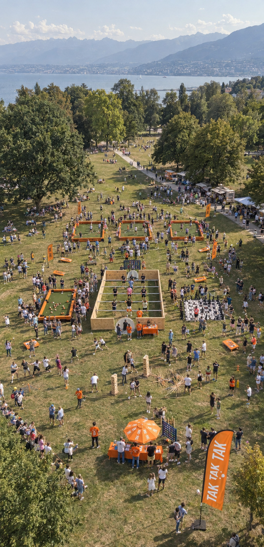 An aerial view of a lively outdoor park event with various game booths, food trucks, and people enjoying activities on a grassy field. There is a large lake and mountains in the background.