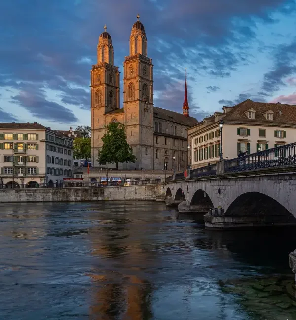 Historical church in Zurich with twin towers over a river, surrounded by older buildings and a stone bridge under a partly cloudy sky at sunset.