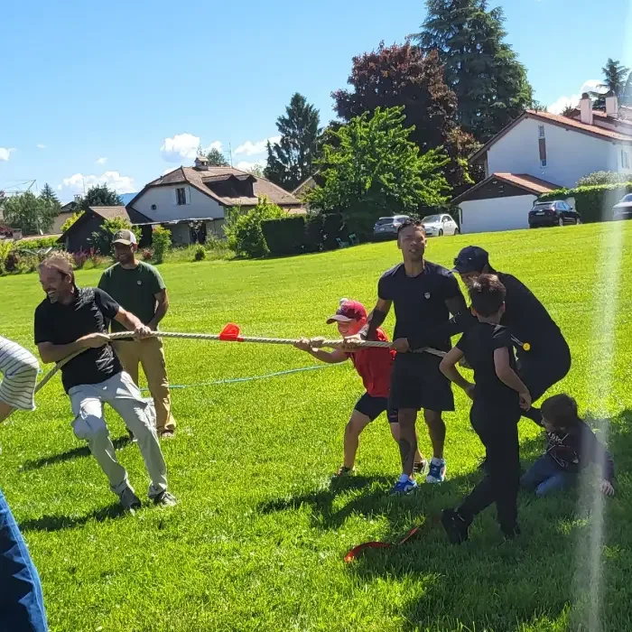 Children and adults are playing a game of tug-of-war on a sunny day in a grassy park, with houses and trees in the background.