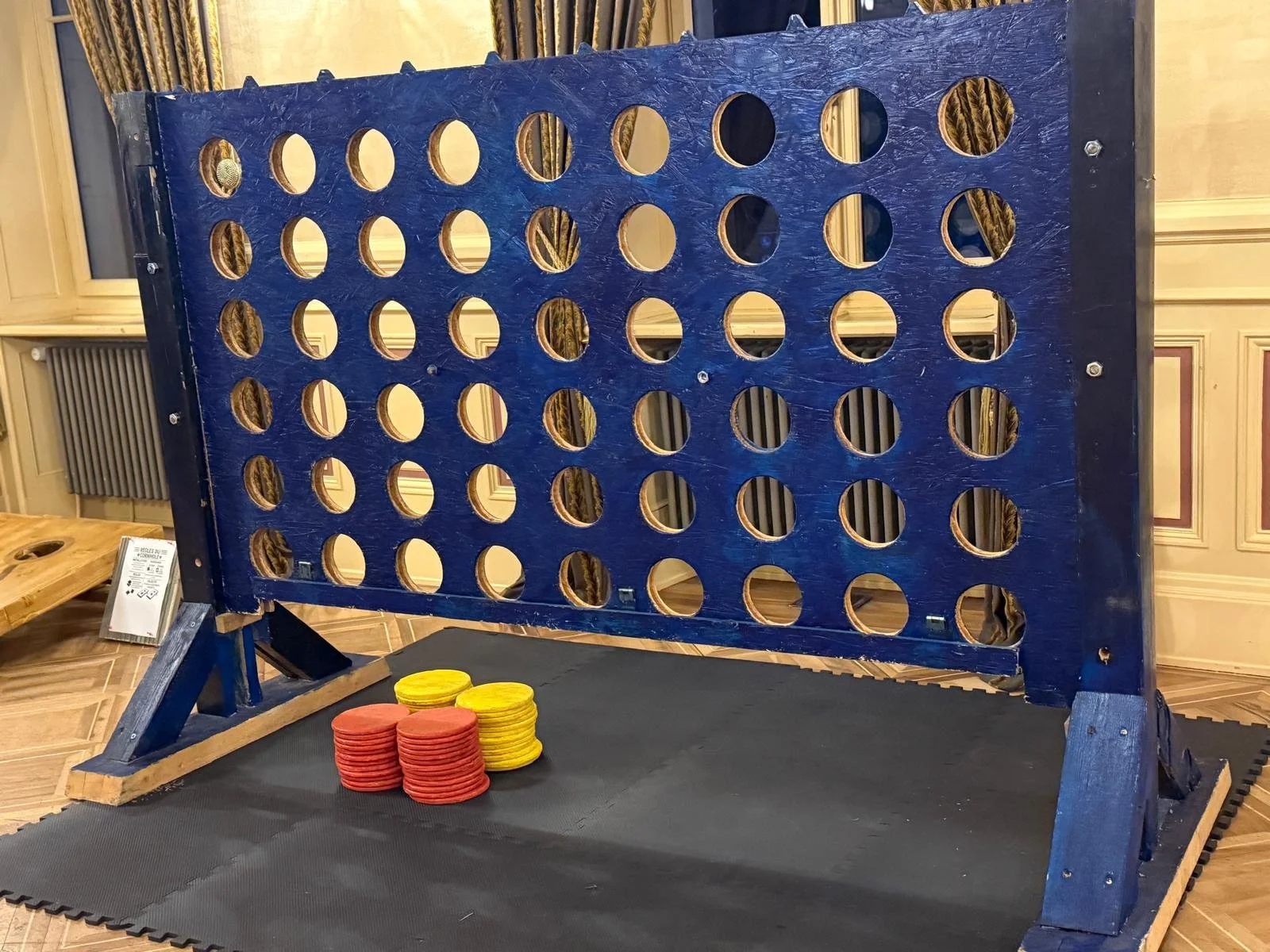 A large wooden connect four game with a blue frame, set up for play in a room with wood-paneled walls and ornate curtains. There are yellow and red chips stacked at the base of the game.