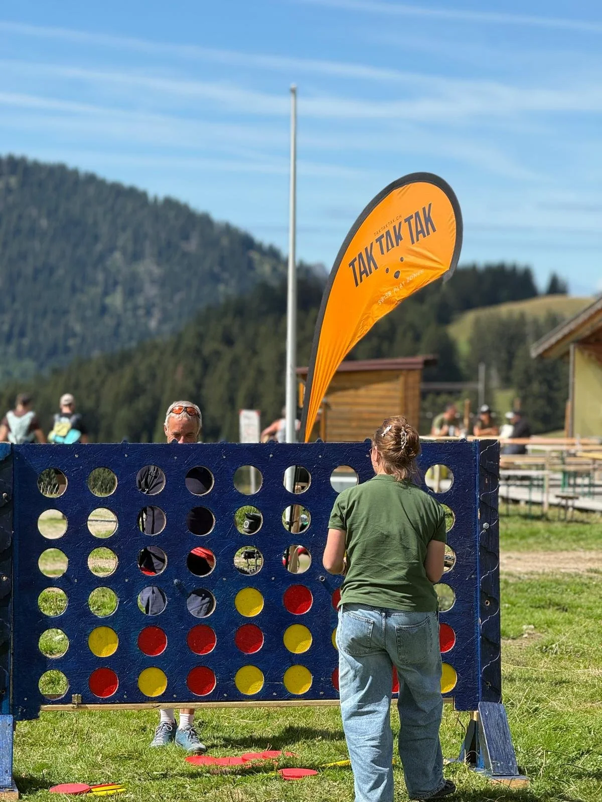 People playing outdoor Connect Four game near wooden buildings with a mountain and blue sky in the background.
