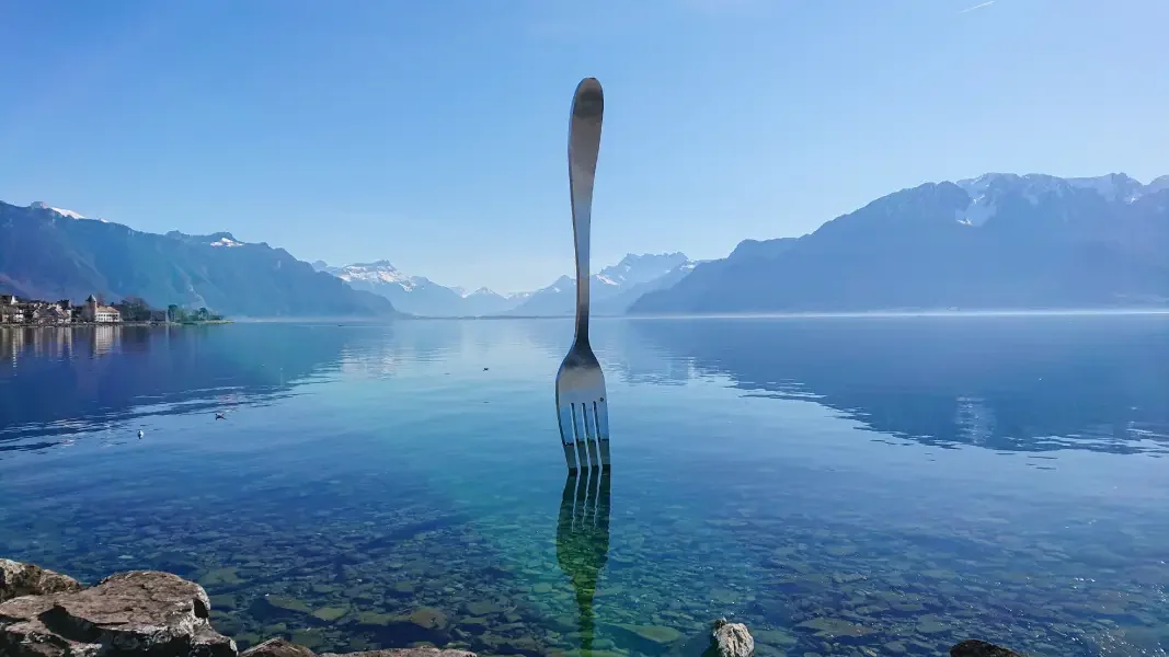 A large fork in Vevey standing upright in a calm lake with mountains in the background and clear blue sky.