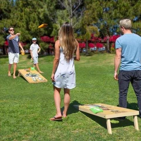 A group of four people playing a bean bag toss game outdoors on a grassy field, with trees and red flowers in the background.