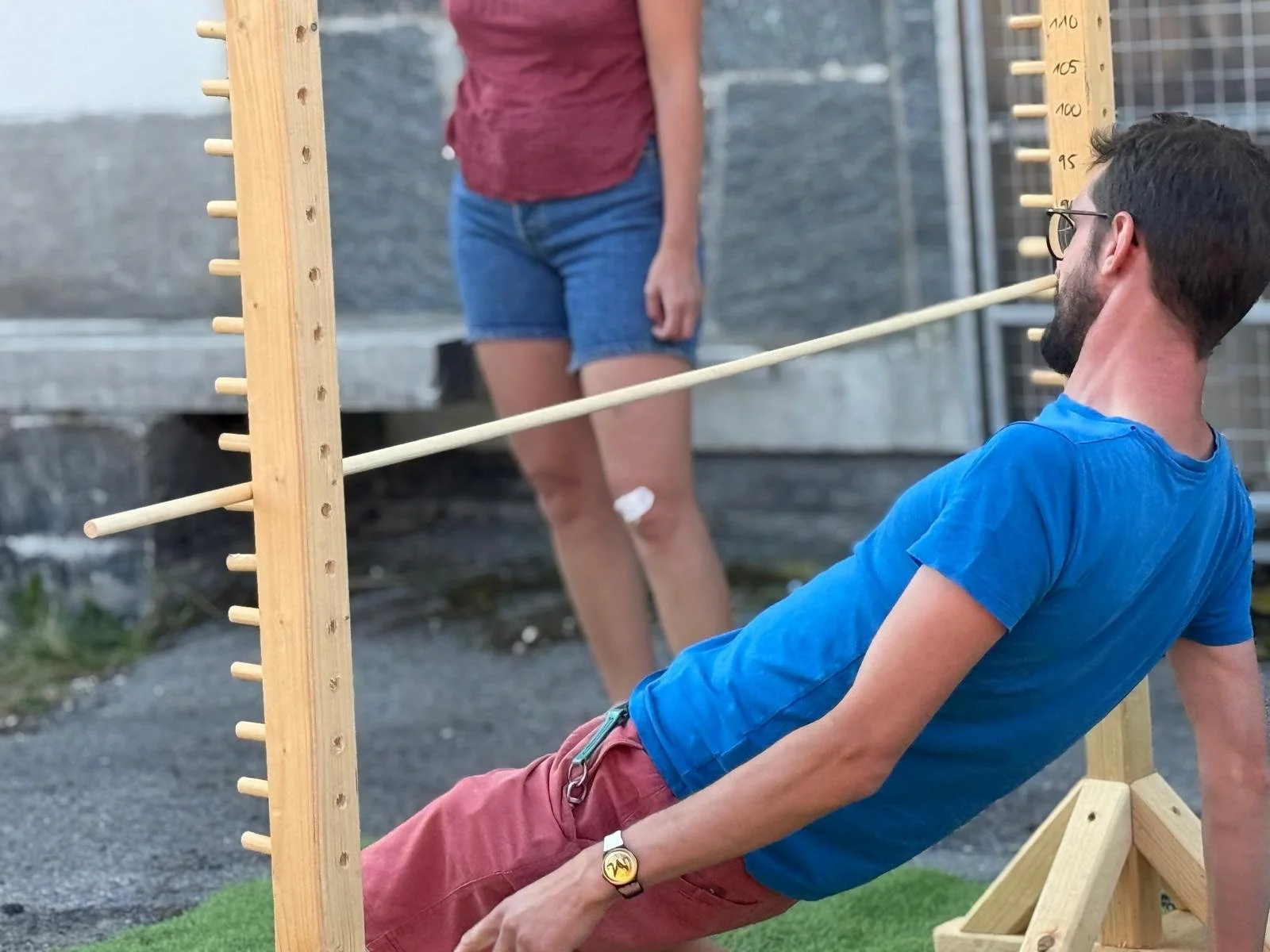 A man in a blue shirt and glasses leaning back on a wooden balance board with a stick through his mouth, while a woman in a red shirt and denim shorts stands behind him, observing. The scene takes place outdoors near a stone wall.