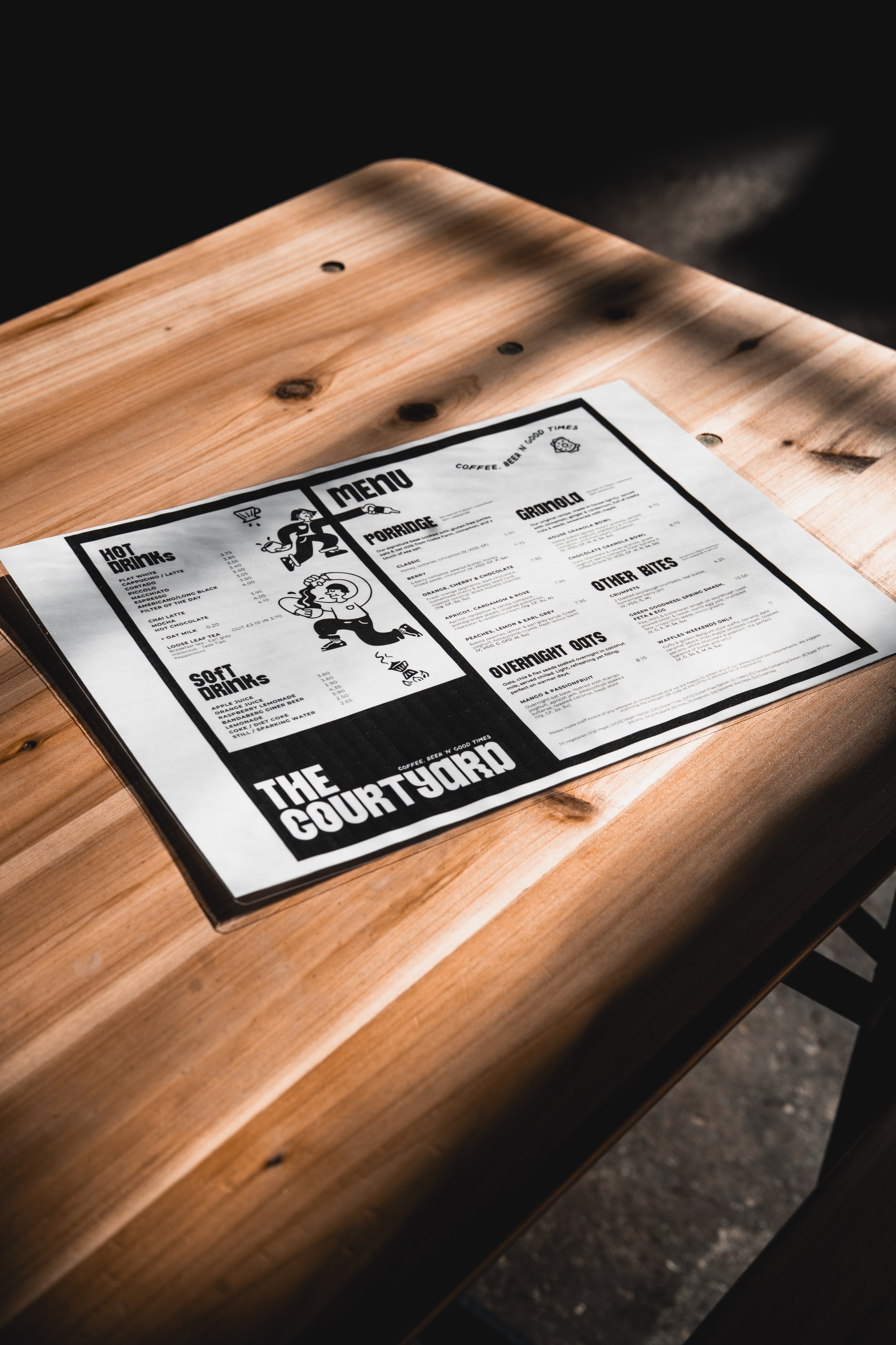 A menu on a wooden table in a cafe or restaurant, featuring sections for hot drinks, soft drinks, porridge, granola, other bites, and overnight oats, with illustrations of animated people.