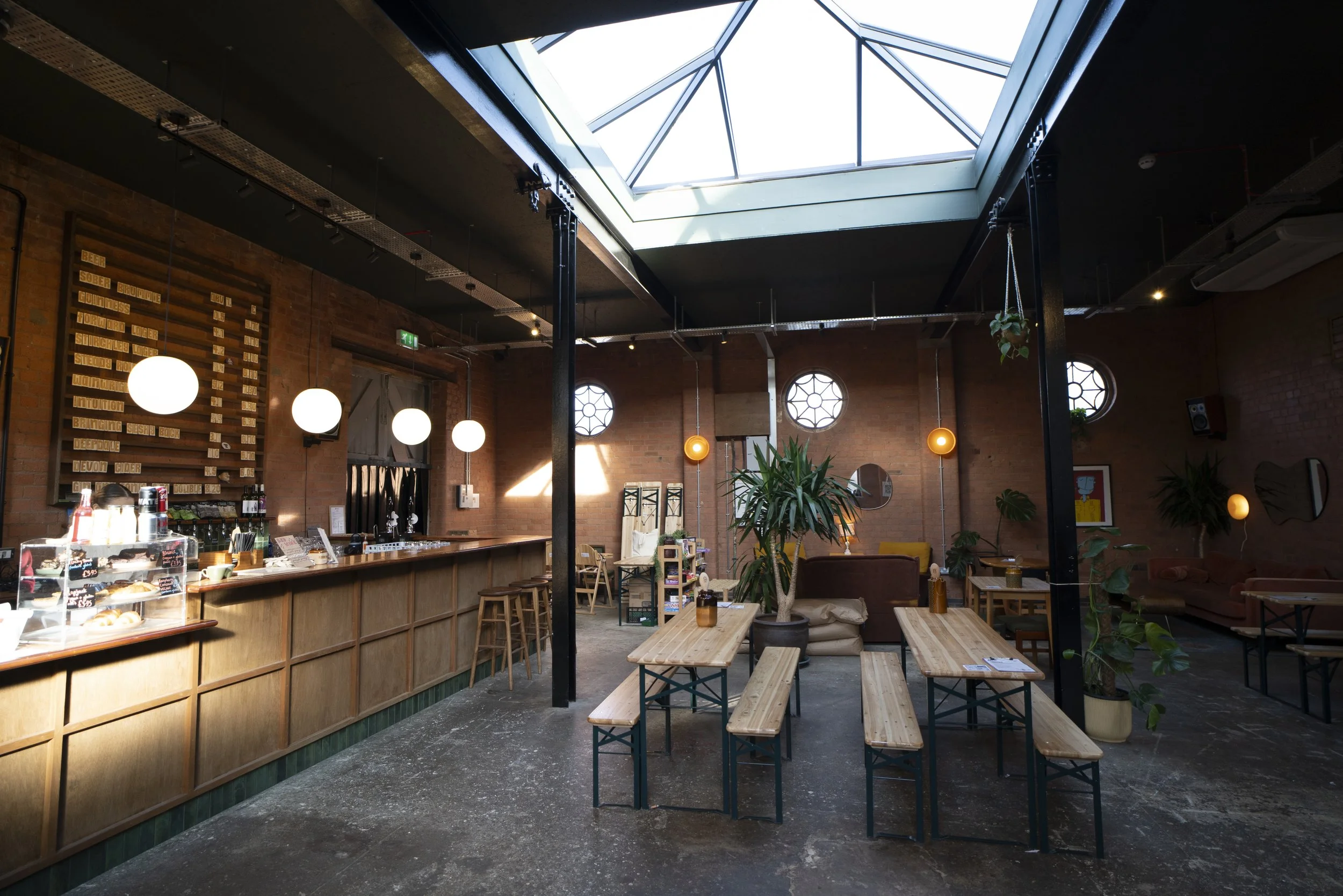 Interior of a cozy cafe or bar with a brick wall, wooden bar counter, hanging spherical lights, and wooden tables and benches. There are large windows allowing natural light, and several potted plants decorate the space.