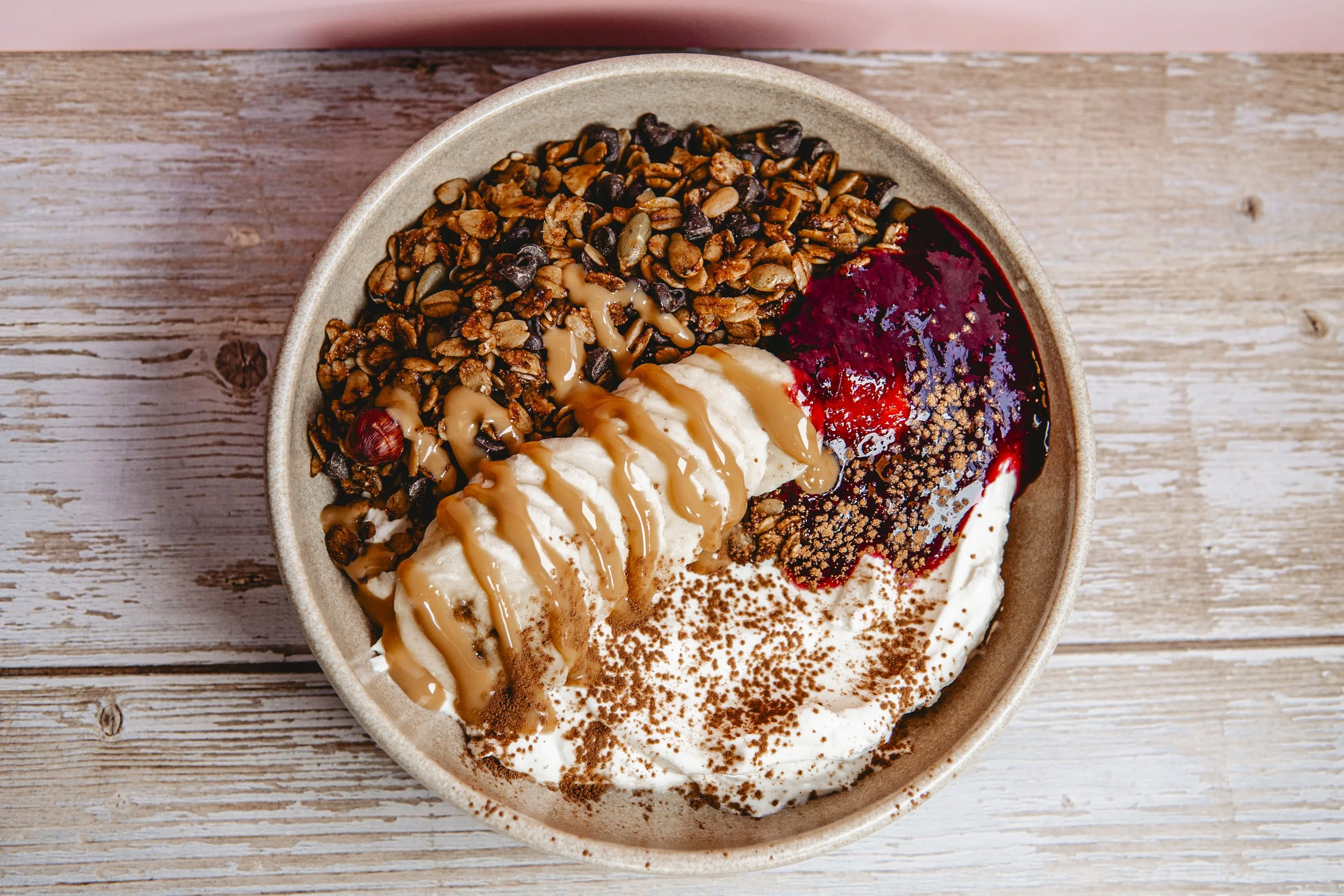 Bowl of frozen yogurt topped with caramel sauce, granola, berry compote, and chocolate sprinkles on a wooden surface.