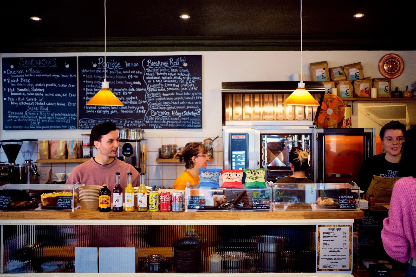 Inside a cozy cafe or bakery, with staff at the counter. Three people are working behind the counter, with food items and drinks on display. A chalkboard menu with breakfast and lunch options is on the wall behind them.
