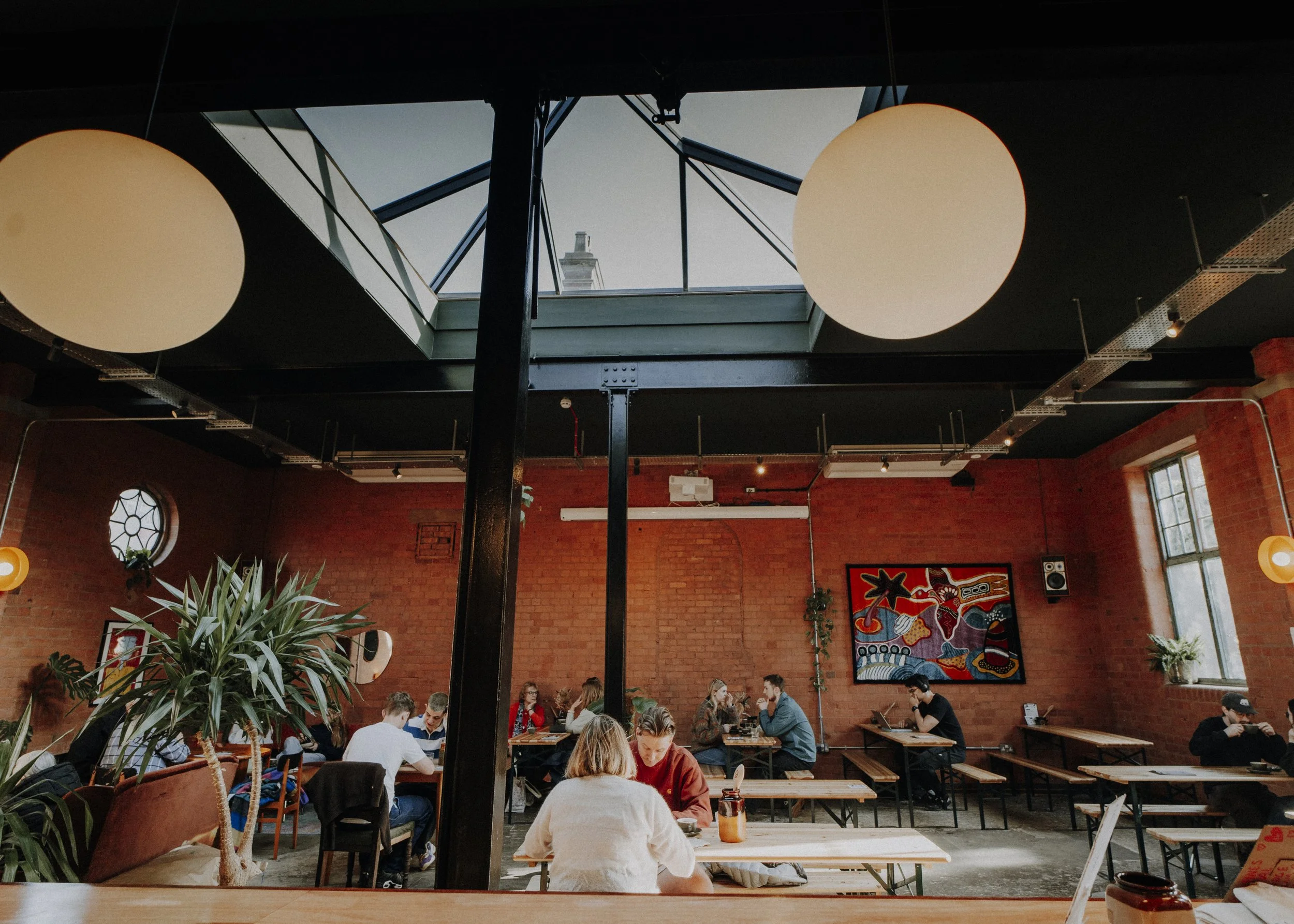 Interior of a modern cafe with exposed brick walls, large windows, hanging circular lights, and a skylight ceiling.