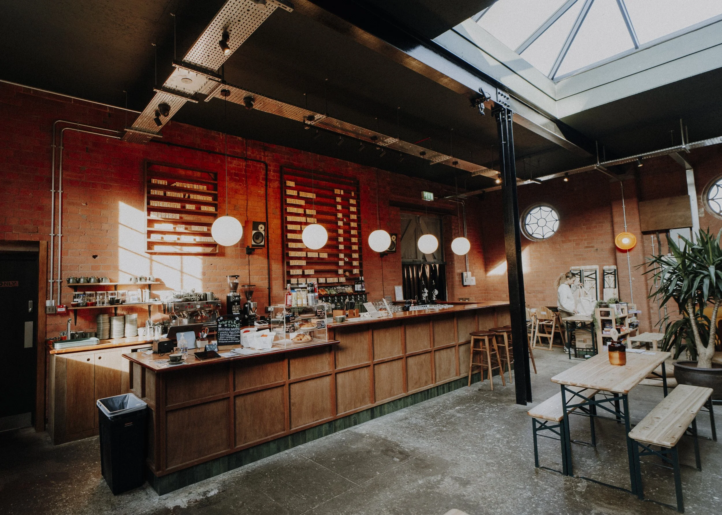 Interior of a cozy cafe with a brick wall, wooden counter, pendant lights, and a seating area with tables and chairs, with a person working in the background.