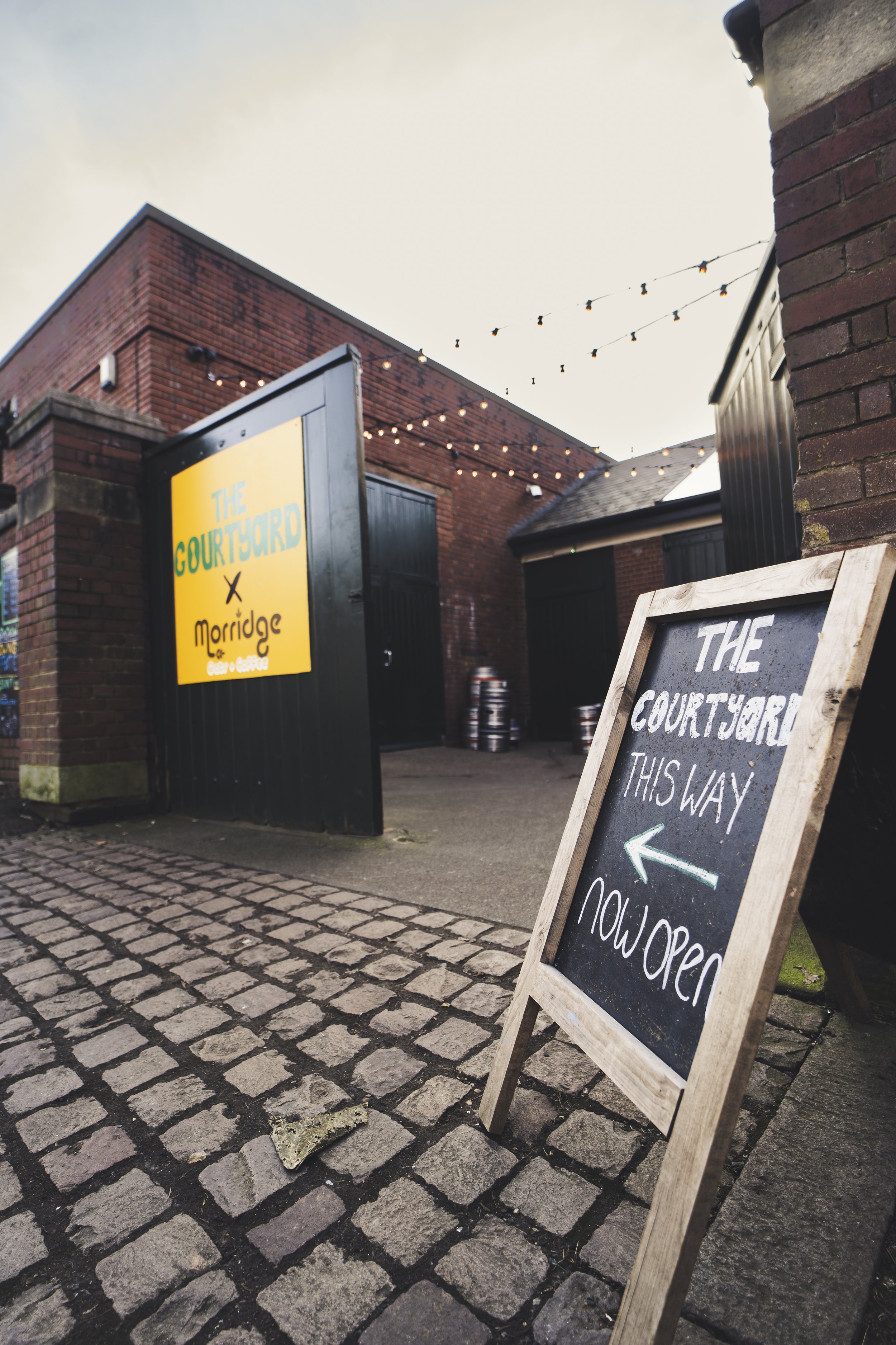 A chalkboard sign outside a brick building directing to the open courtyard for a venue called Morridge, with string lights overhead and a cobblestone pavement in the foreground.