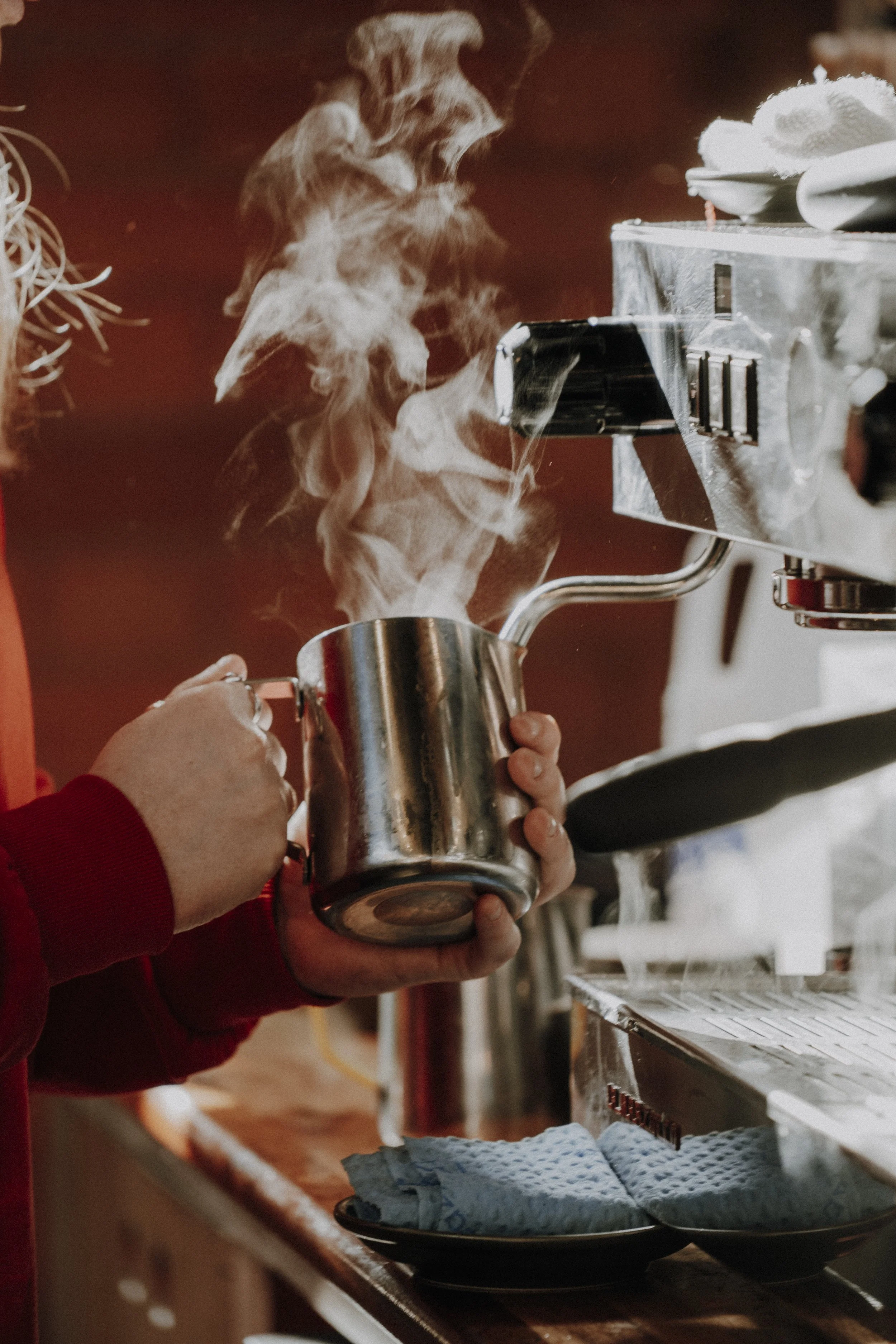A person in a red shirt is steaming milk in a stainless steel pitcher under an espresso machine.
