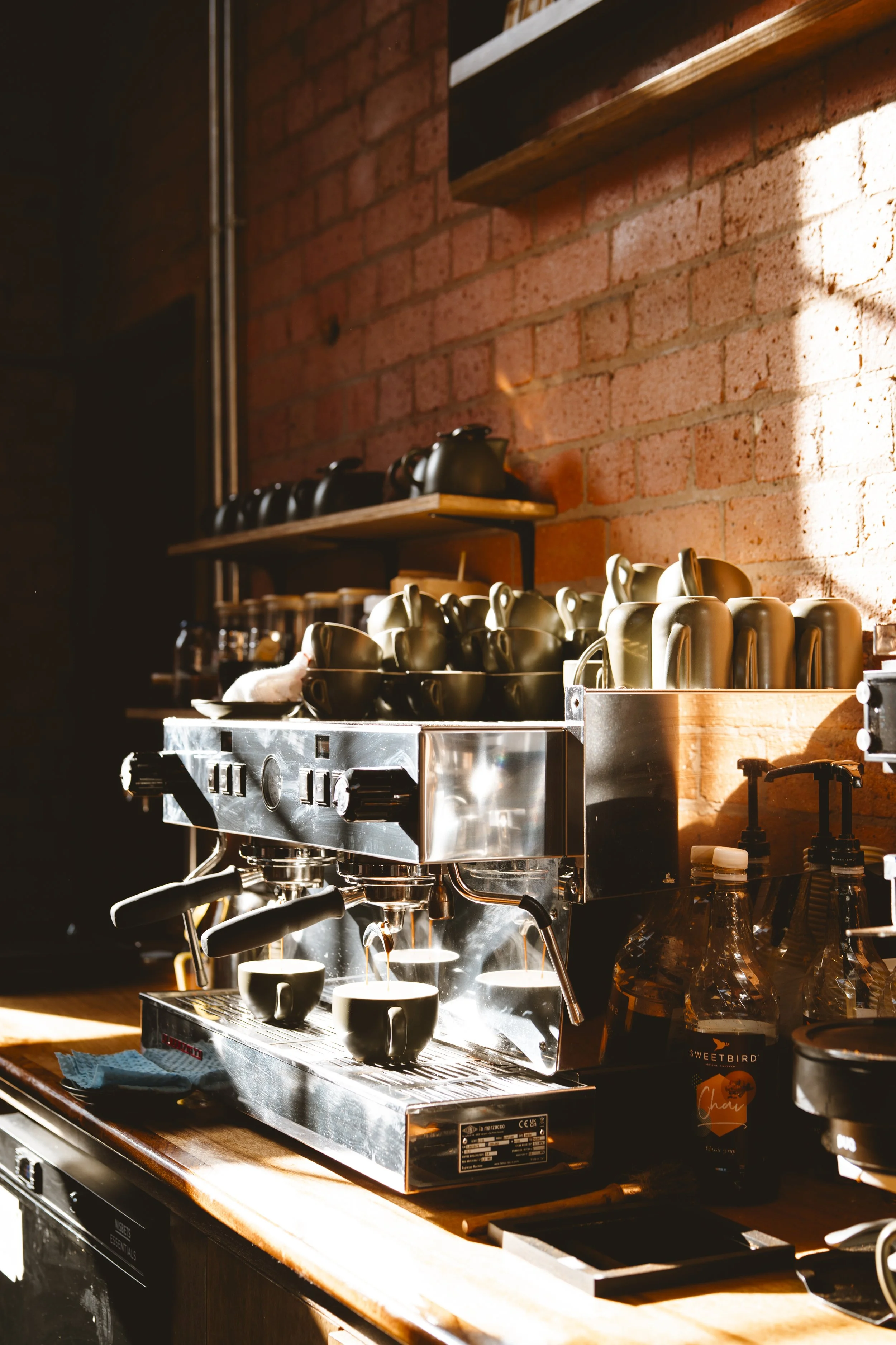 Close-up of a commercial espresso machine brewing coffee into cups, with shelves of mugs and a brick wall in the background.