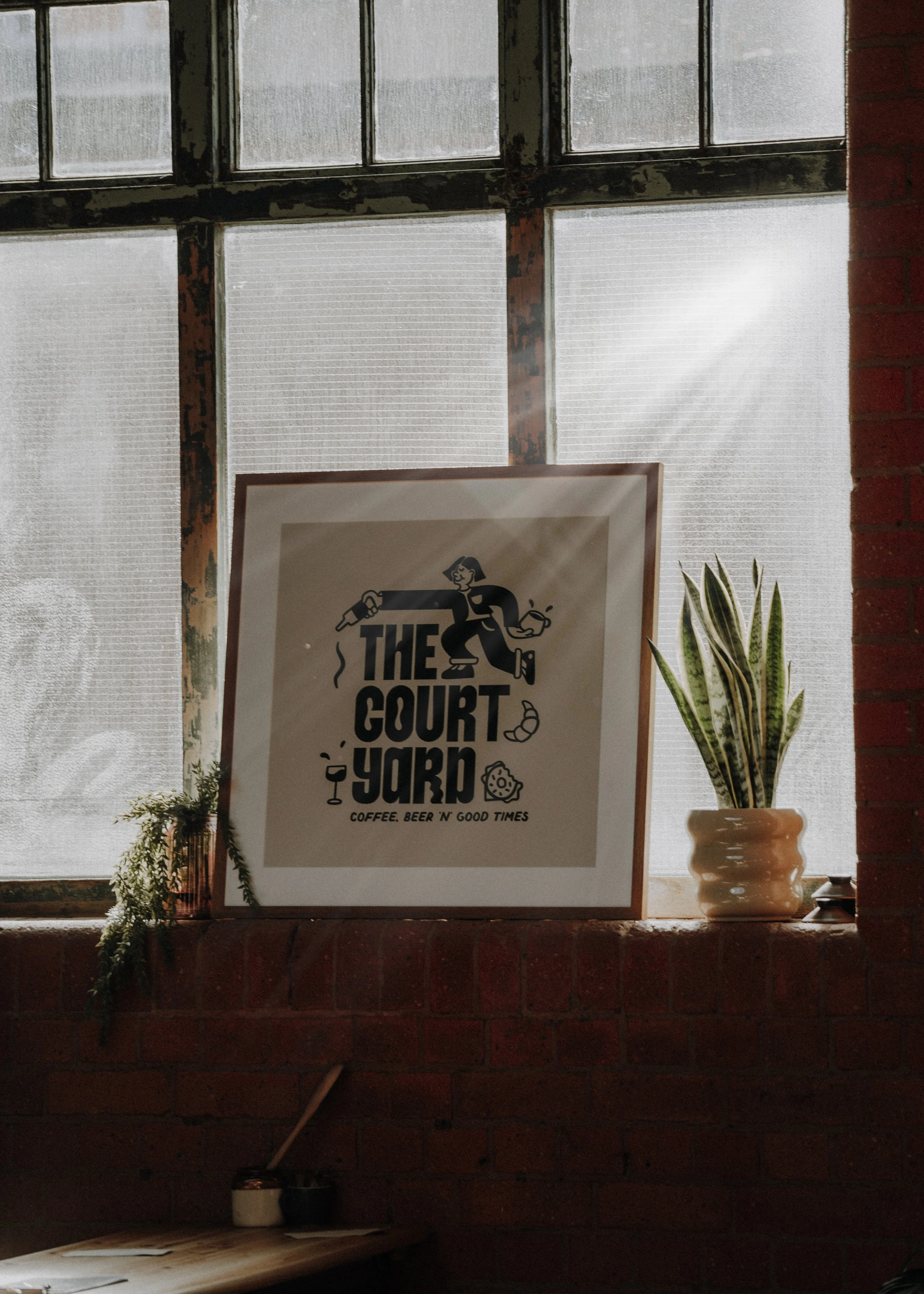 Sign in a frame on a windowsill reading "The Court Yard" with illustrations of a person holding a paint roller, a wine glass, and a piece of cheese. The window behind has frosted glass panes, and there are potted plants beside the sign.