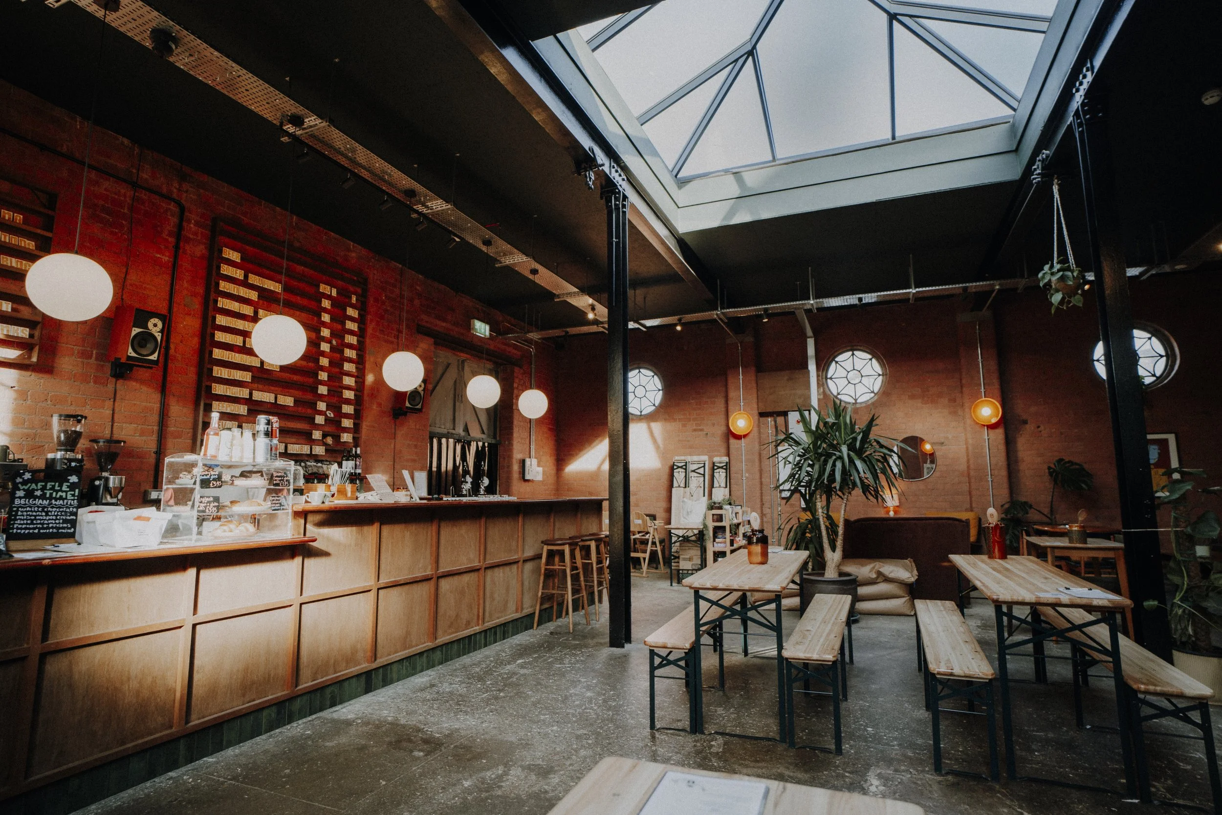 Interior of a modern cafe with brick walls, hanging globe lights, wooden tables and benches, large skylight, and potted plants.