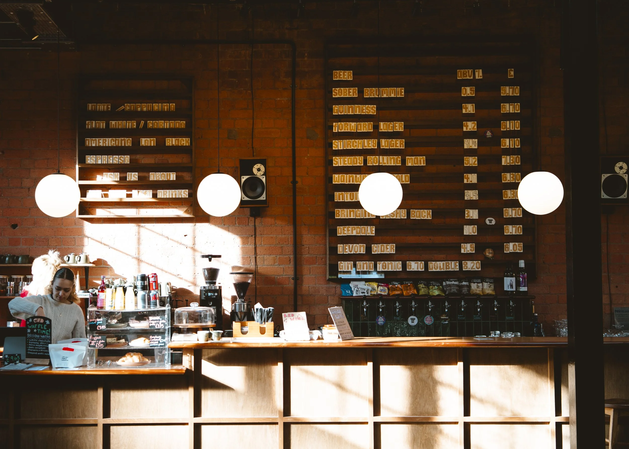 Inside a coffee shop with a brick wall, three round white pendant lights, a menu board with drink names and prices, a barista behind the counter, and various coffee-making equipment.
