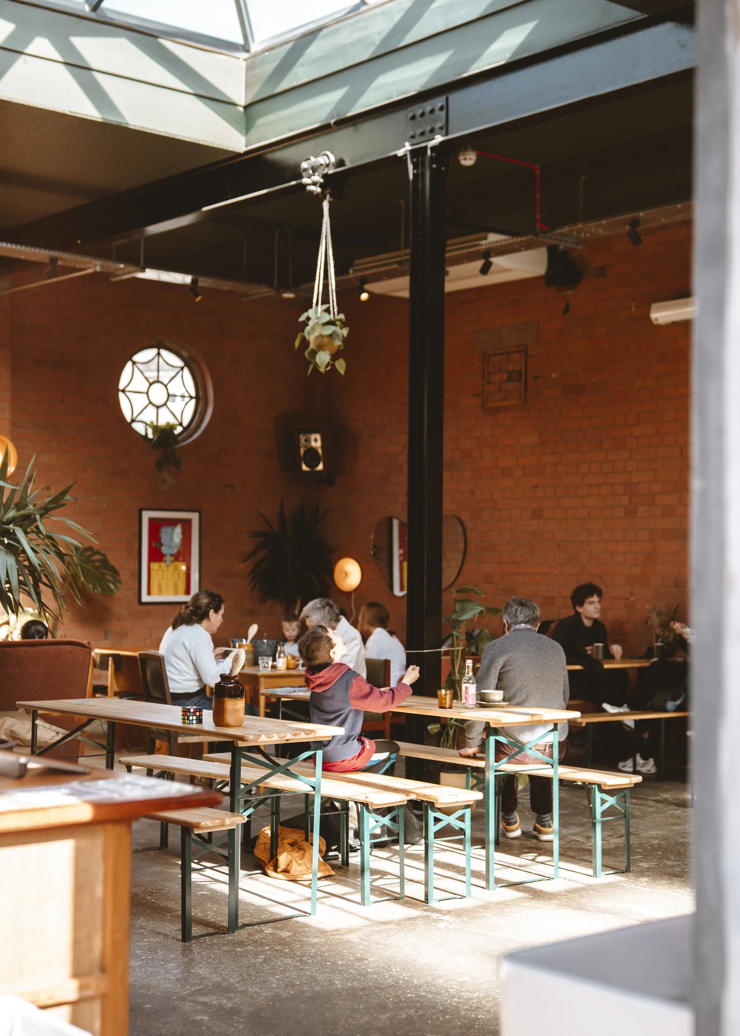 People sitting at wooden tables and benches inside a cafe with brick walls and artwork, some drinking, some working on laptops.