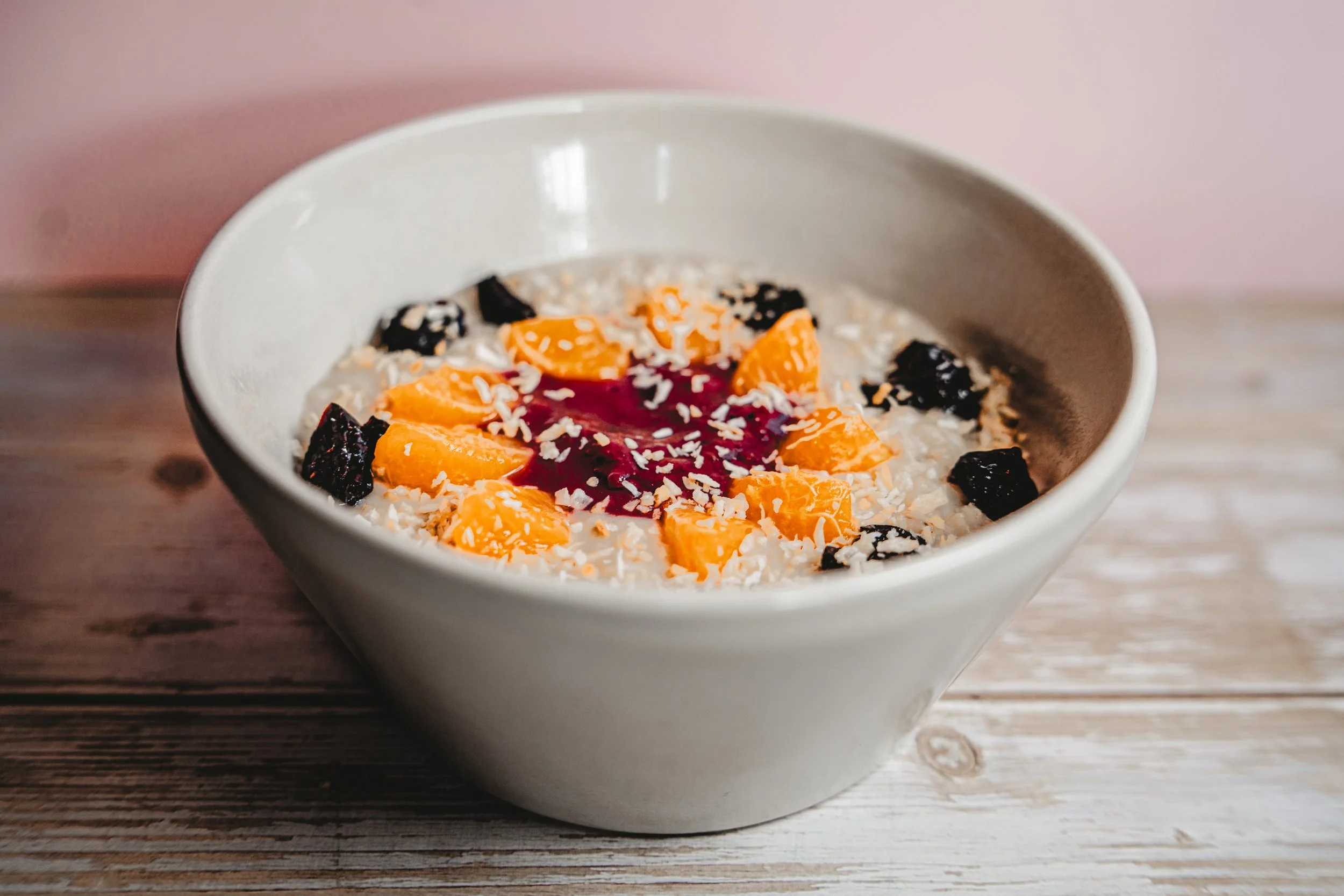 A bowl of oatmeal topped with orange slices, dark berries, shredded coconut, and a drizzle of berry sauce on top, placed on a wooden surface.