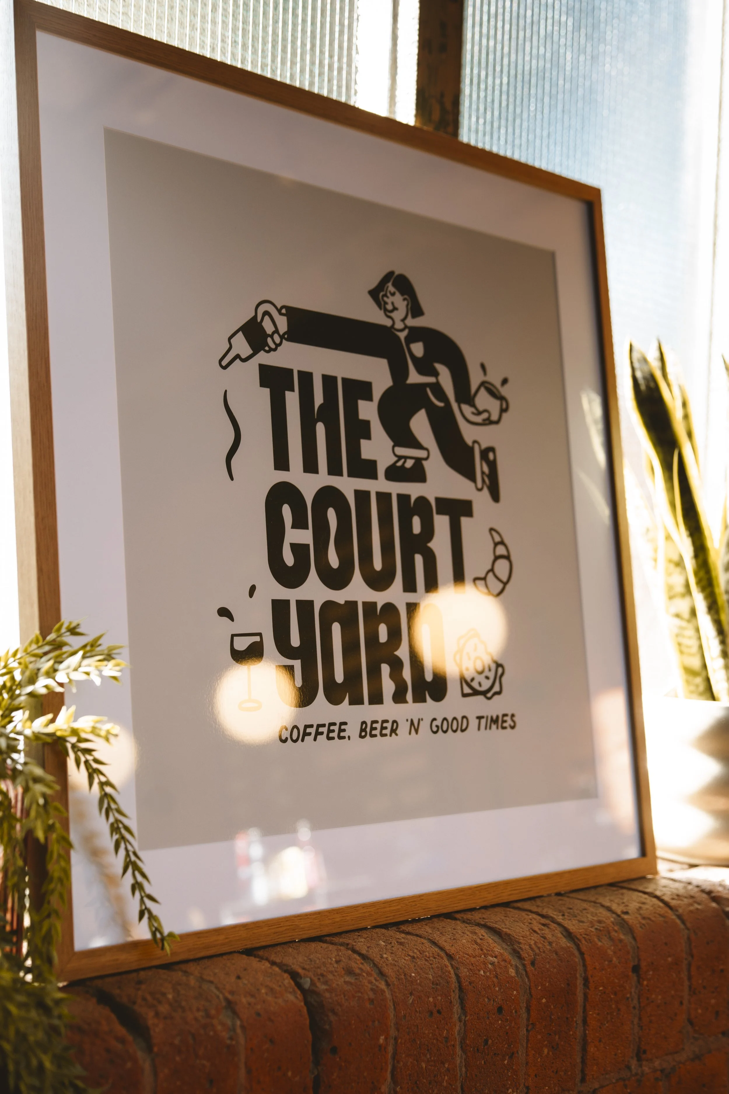 A framed poster on a brick ledge with the text 'The Court Yo' and a drawing of a person holding a cup, along with smaller text 'Coffee, Beer 'N' Good Times'.