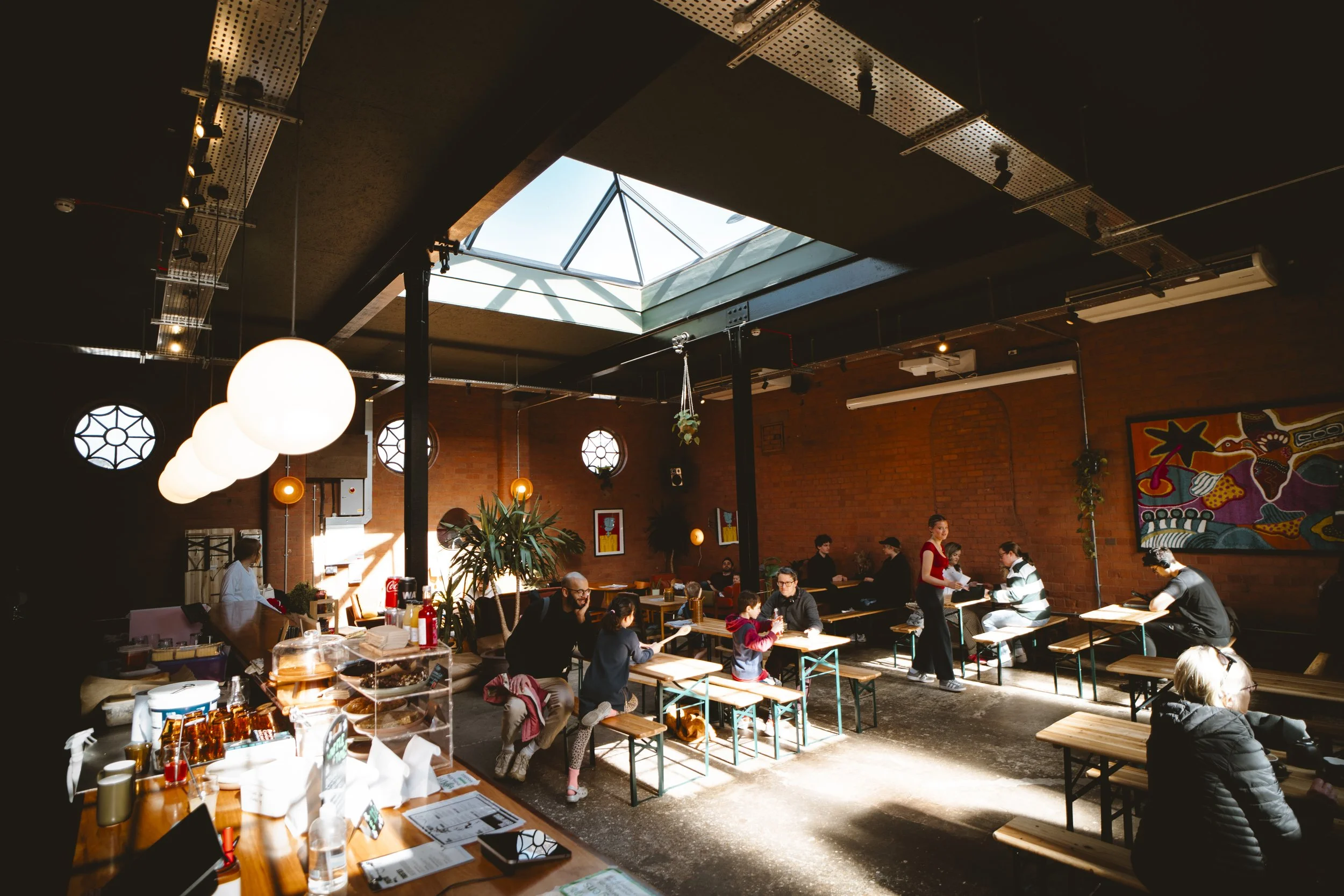 People dining in a sunlit indoor restaurant with brick walls, a skylight, hanging lights, and colorful artwork.