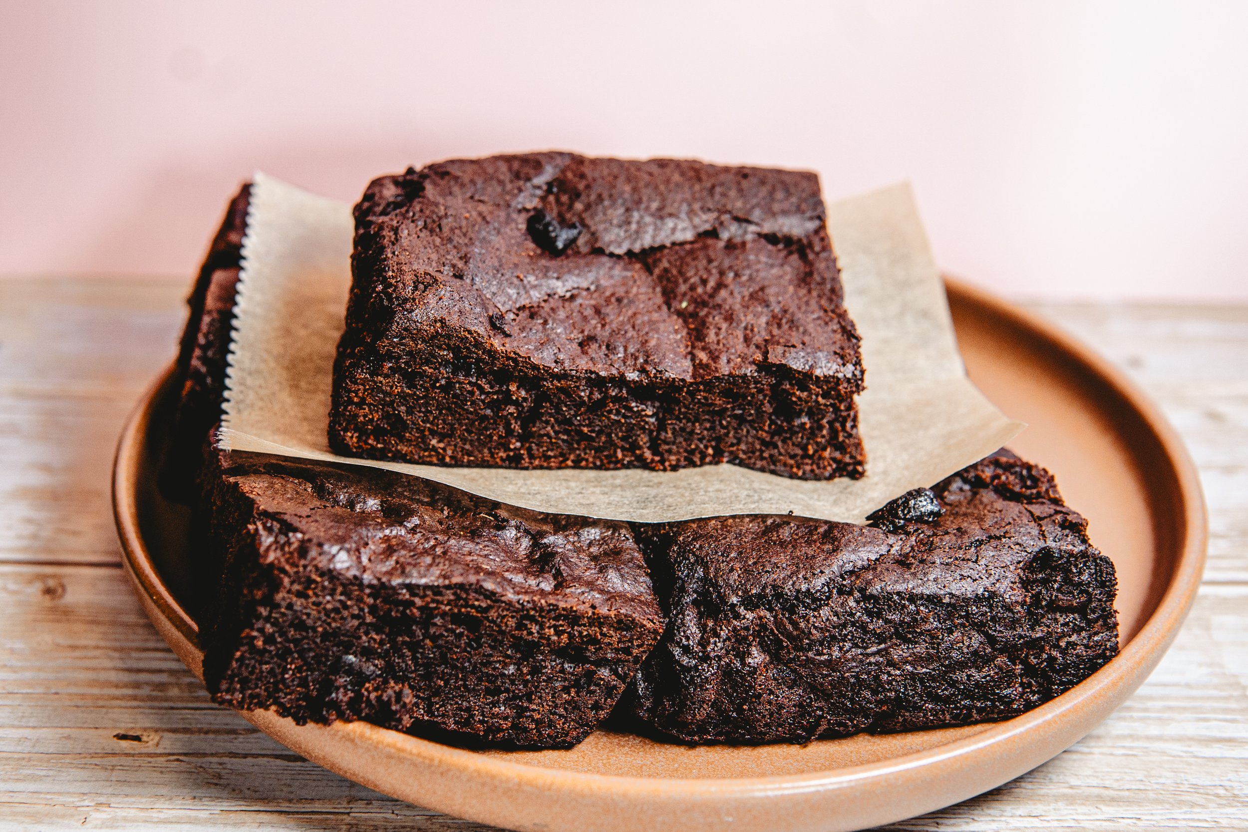 Three square chocolate brownies on a brown plate with parchment paper, placed on a wooden surface.