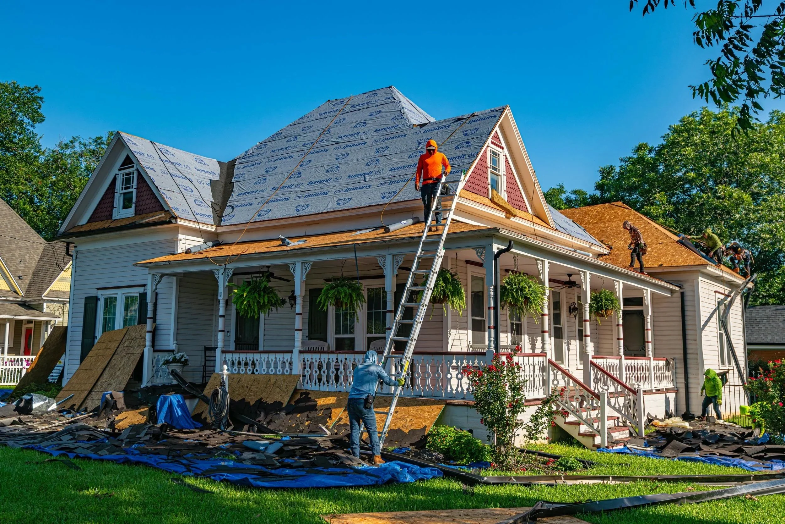 People working on a house roof renovation, installing new roofing shingles, with some workers on the roof and others below. The house has a front porch with hanging plants and is surrounded by a green lawn and trees.