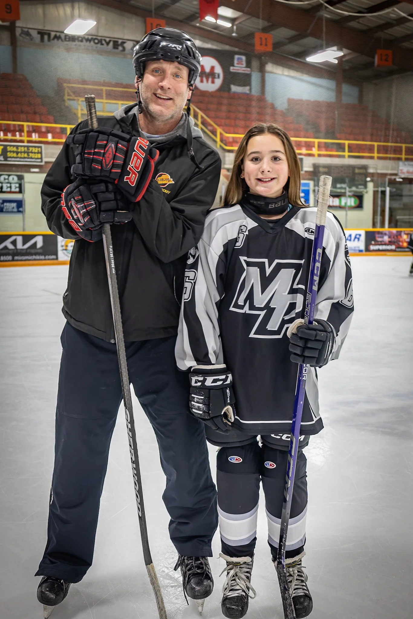 A man and a young girl in hockey gear standing on an ice rink, smiling for the photo, with hockey sticks in their hands.
