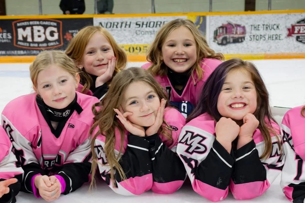 Five young girls in pink and black hockey jerseys lying on the ice rink, smiling at the camera.