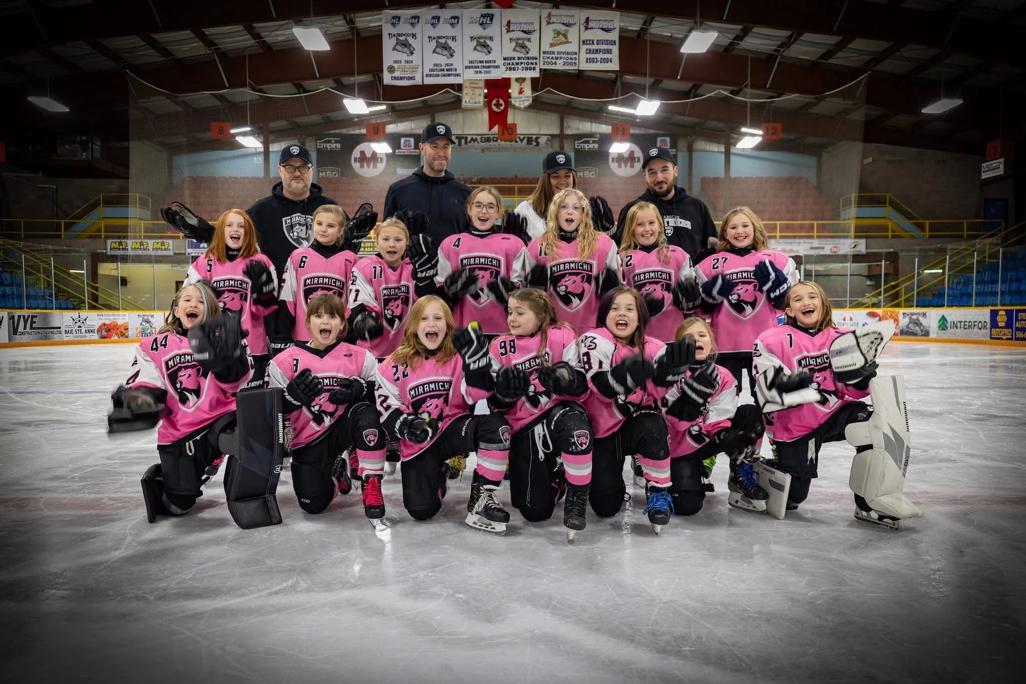 A youth hockey team in pink and white jerseys posing with their coaches on an ice rink, celebrating after a game.