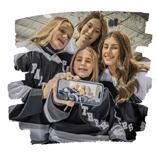 Four young women in hockey jerseys taking a selfie together at an indoor ice rink.