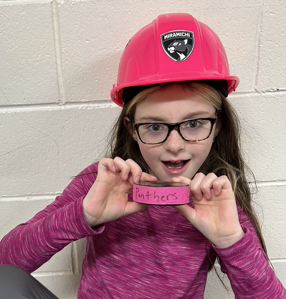 A girl wearing glasses and a pink hard hat holding a pink wristband with the word 'Panthers' written on it, standing against a beige wall.