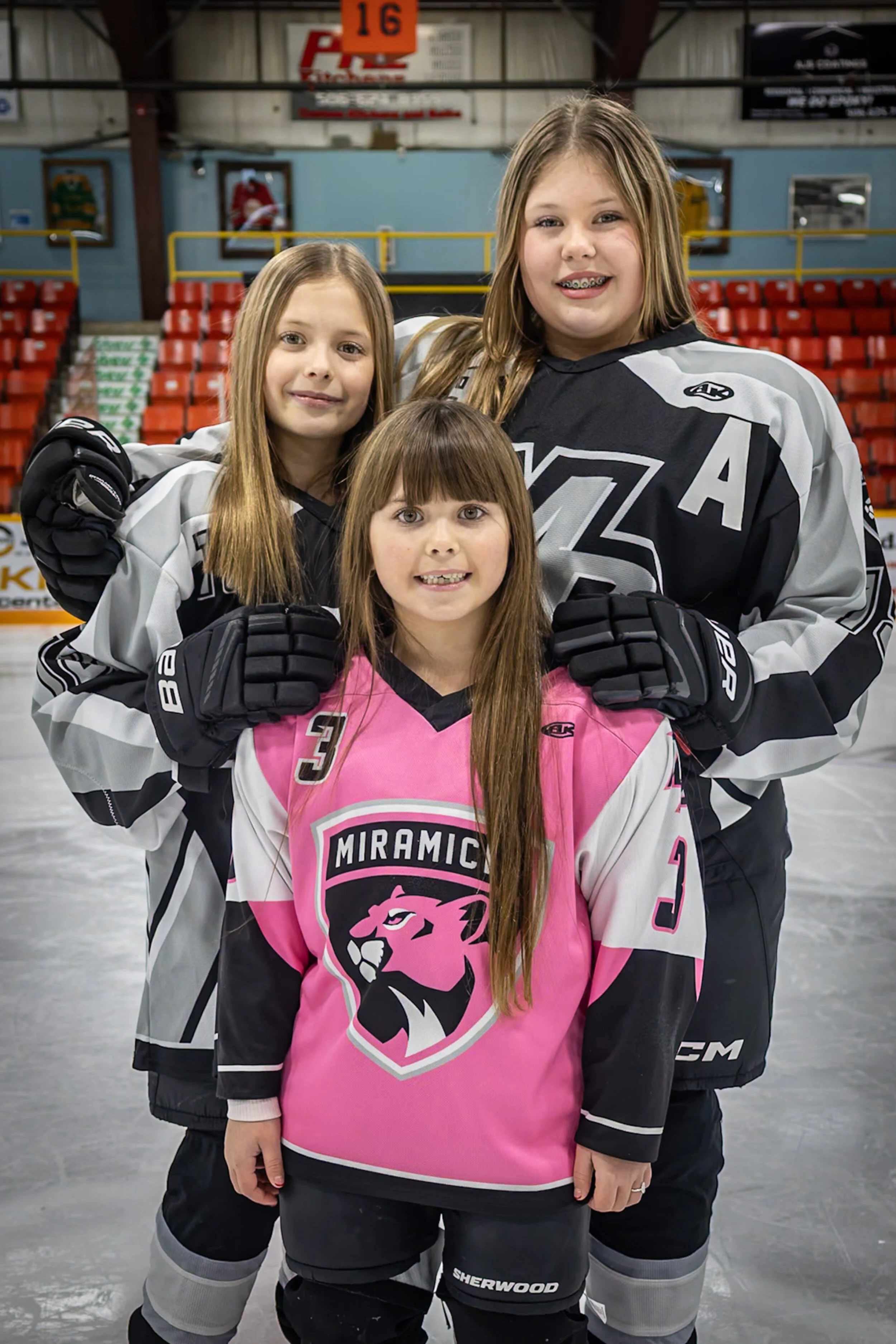 Three young girls in hockey jerseys standing on an ice rink, smiling at the camera, with seating and signs in the background.