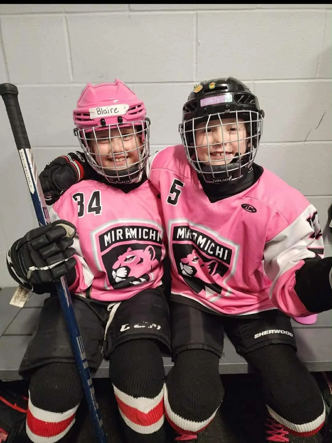 Two young ice hockey players in pink jerseys sitting on a bench, smiling, wearing protective gear and black helmets, with their arms around each other.