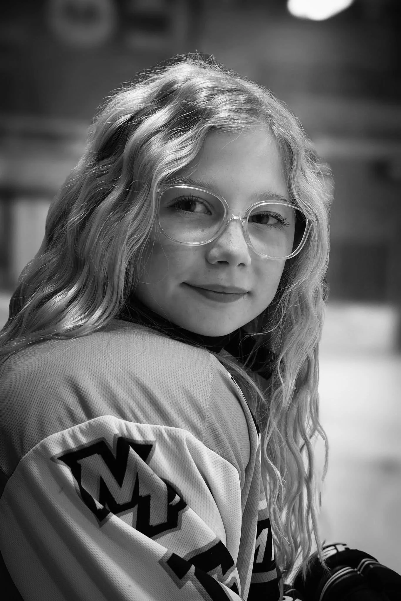 A young girl with long, wavy hair wearing large round glasses and a sports jersey with a patch on the sleeve, looking over her shoulder with a slight smile.