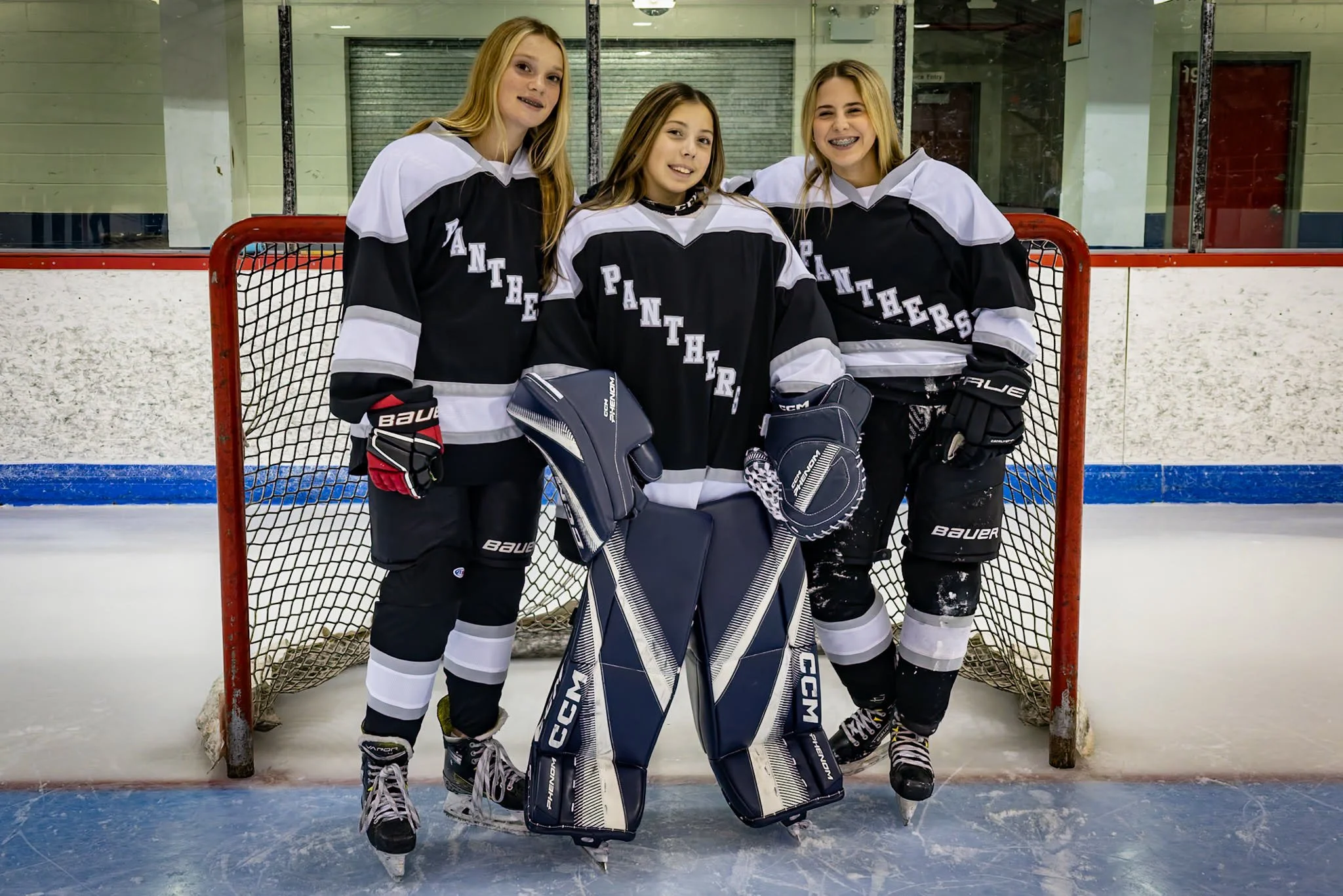 Three young female ice hockey players in black and white jerseys with 'Panthers' written on them, standing on ice rink in front of goal net, wearing hockey equipment including skates, gloves, and pads, smiling.