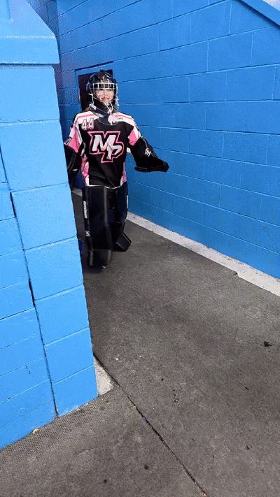 A young girl in hockey gear, including a helmet, jersey, and leg pads, peeking out from behind a blue brick wall in an indoor ice hockey arena.