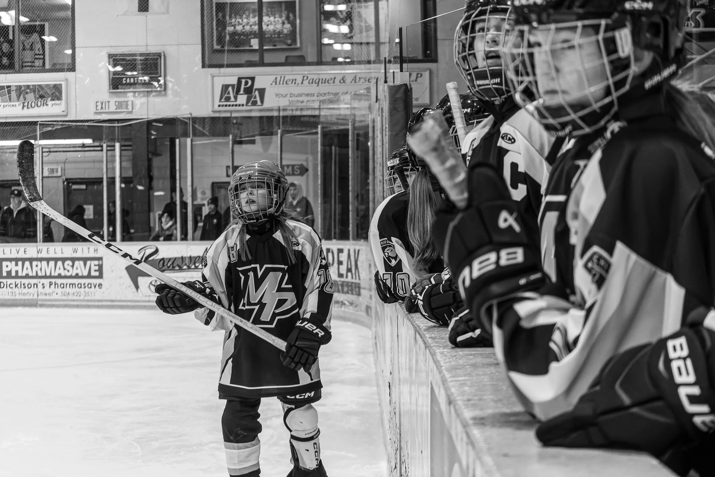 A young girl ice hockey player in uniform standing on the ice rink, holding her hockey stick, while her teammates are sitting on the bench behind her.