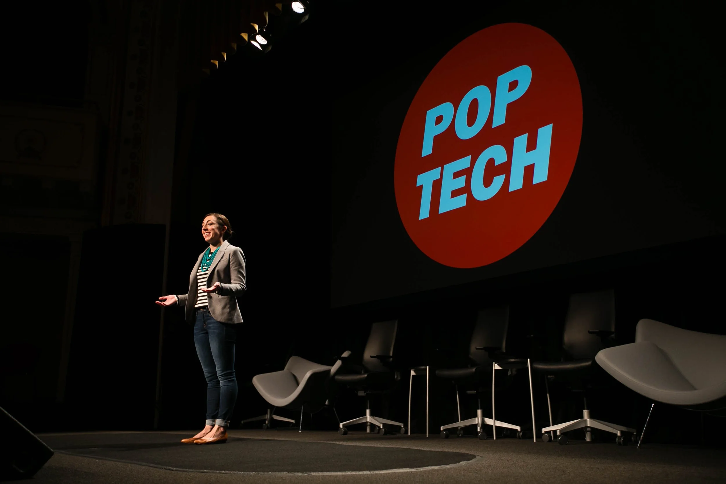 Jessica Lawrence Quinn giving a presentation on stage with a large screen behind her displaying the words "Pop Tech" inside a red circle.