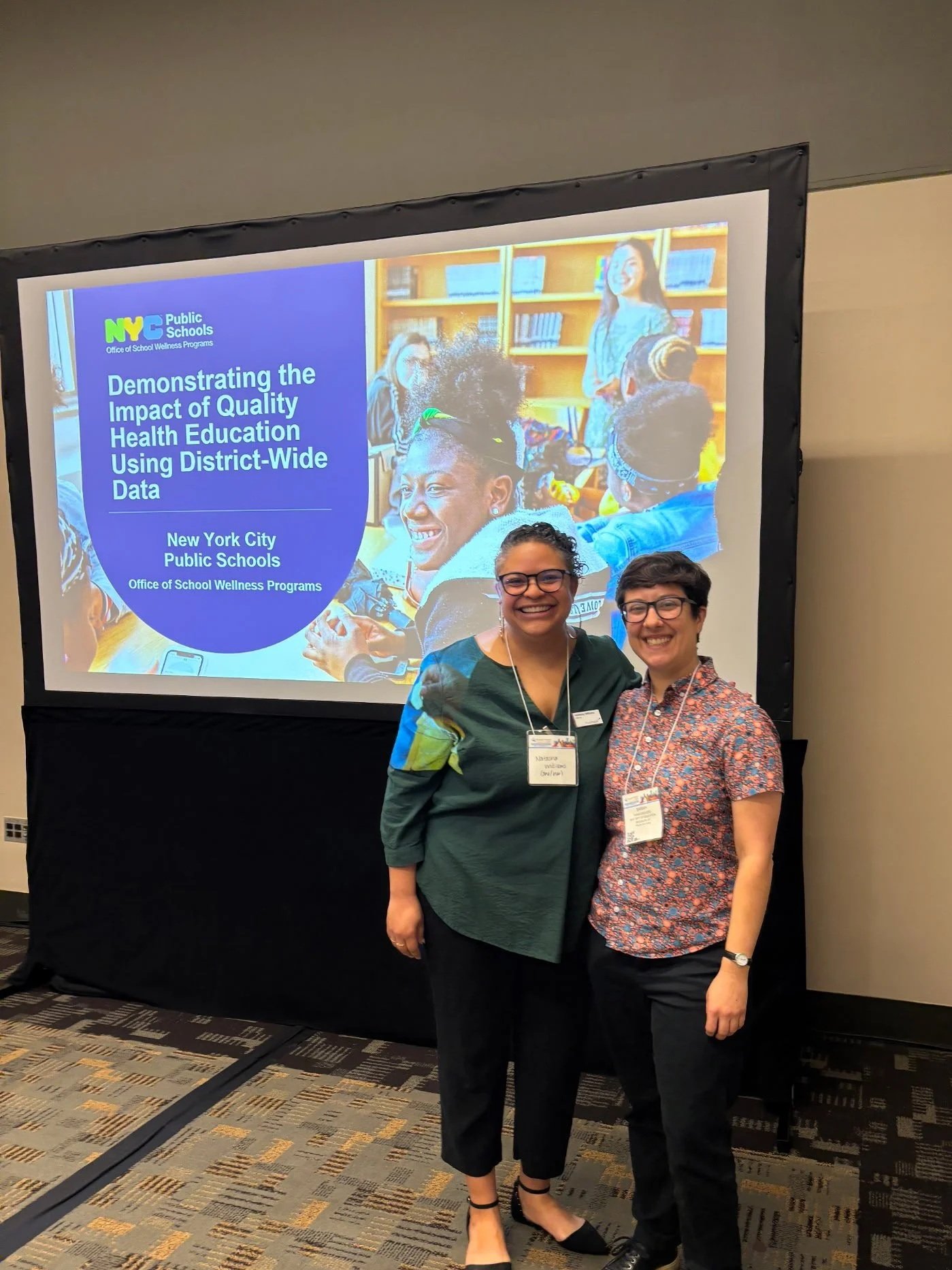 Two women smiling and standing in front of a presentation screen at a conference. The screen displays a slide related to New York City Public Schools and health education.