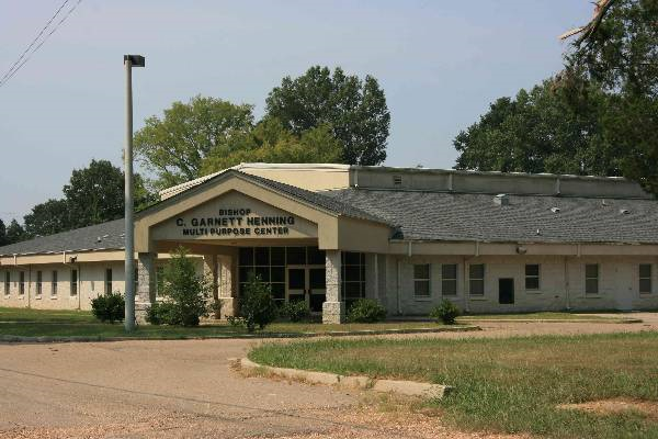 A single-story building labeled 'DEMO C. GARNETT HENNING MULTI PURPOSE CENTER' in a rural area with a grass lawn and trees in the background.