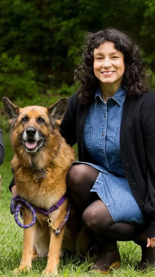 Woman with curly dark hair smiling next to a friendly, happy German Shepherd dog in a park.