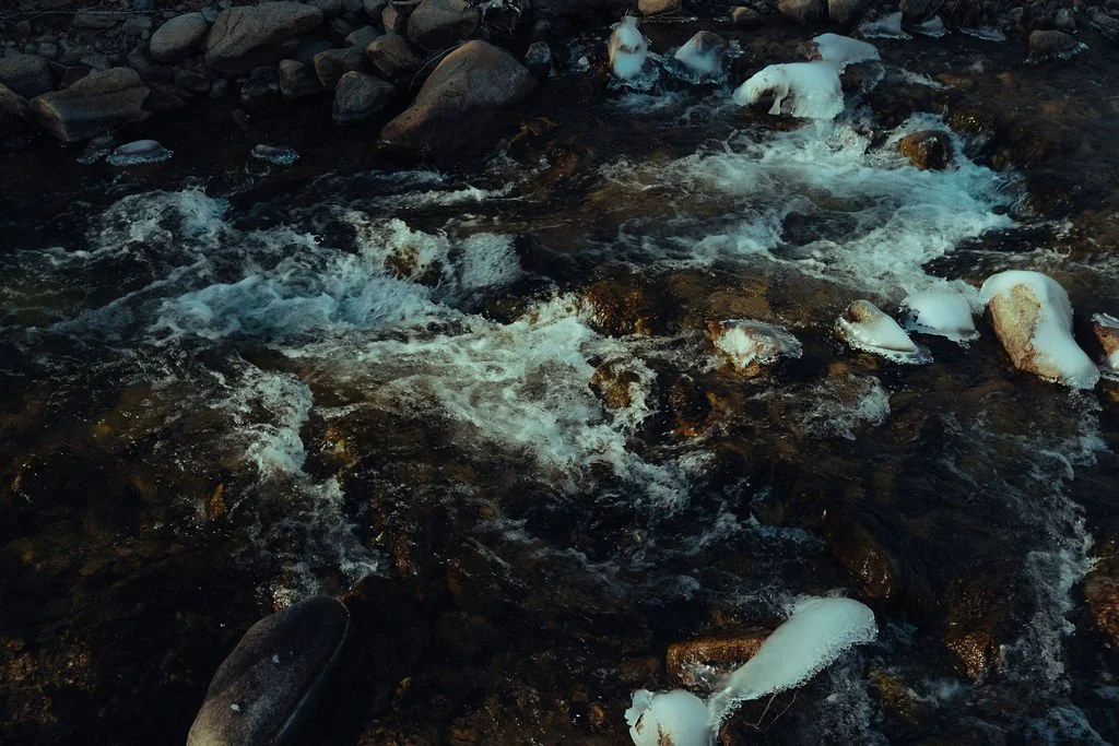 A flowing mountain stream with rocks and patches of snow.