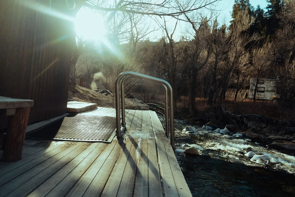 A wooden dock with metal handrails extending over a stream, with trees in the background and sunlight shining through.
