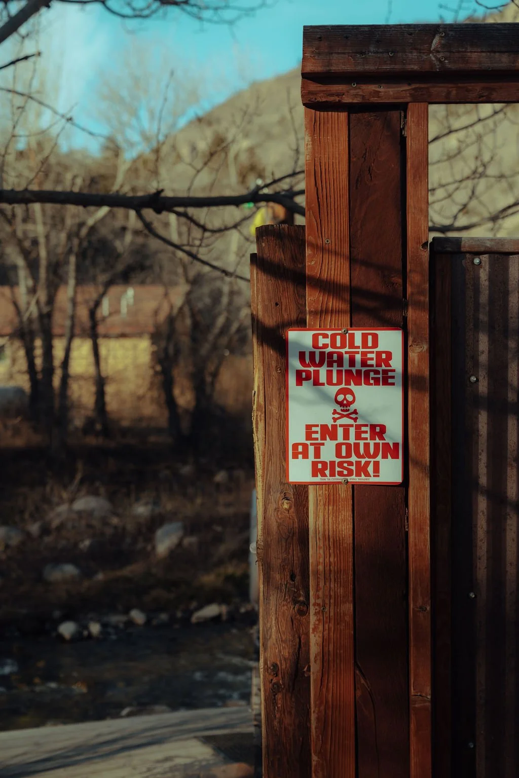 A warning sign on a wooden fence reads 'Cold Water Plunge, Enter at Own Risk' with a skull and crossbones graphic, in an outdoor setting with leafless trees and rocky ground.