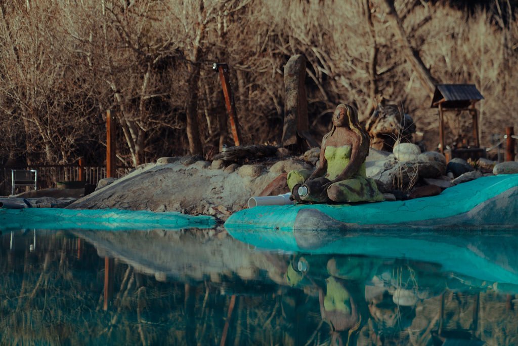 A decorative scene with a sitting woman sculpture by a pool, rocks, leafless trees, and small structures in the background.