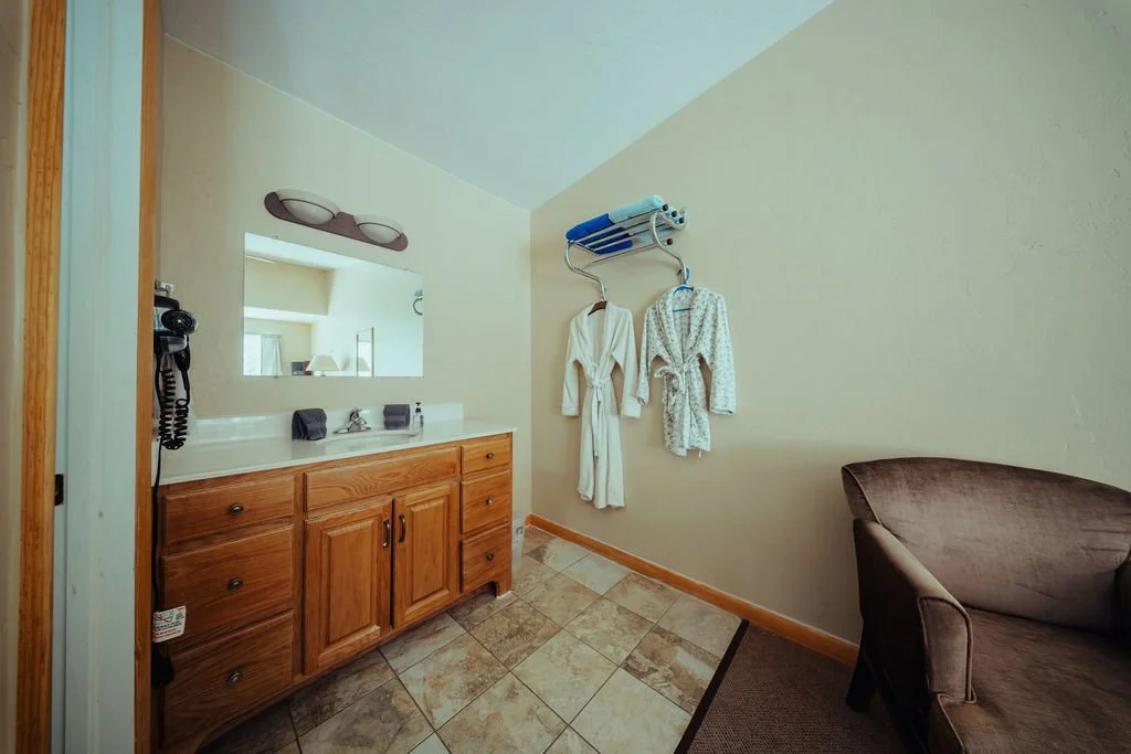Hotel room with a wooden vanity, mirror, wall-mounted hairdryer, bathrobes hanging on a wall rack, towels, a brown armchair, and tiled flooring.