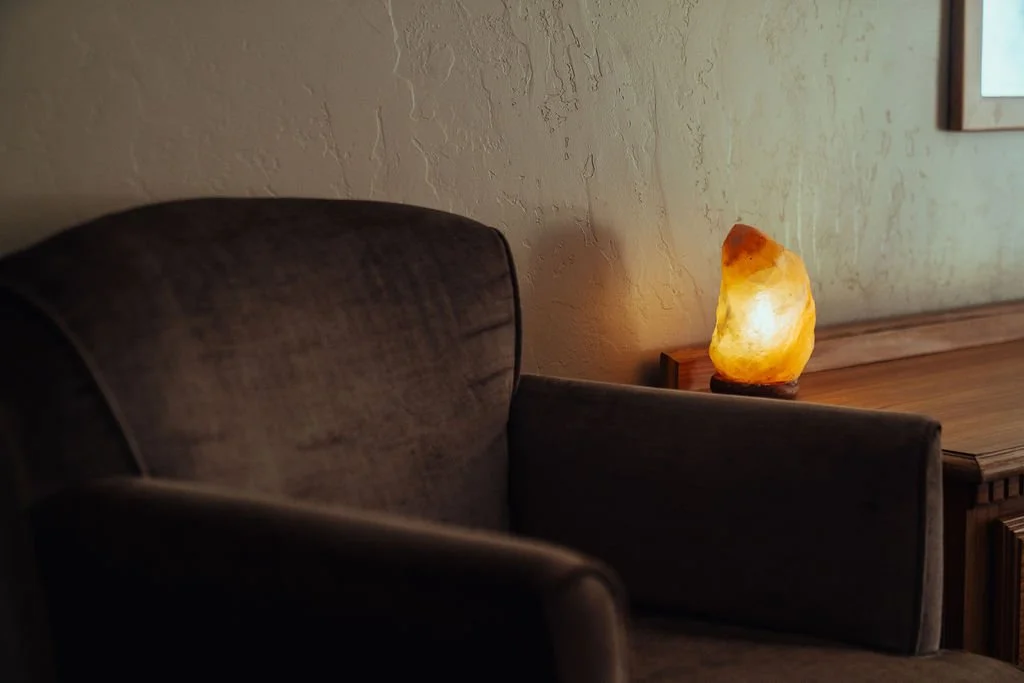 A salt lamp on a wooden table next to a wall, with part of a dark upholstered chair visible in the foreground.