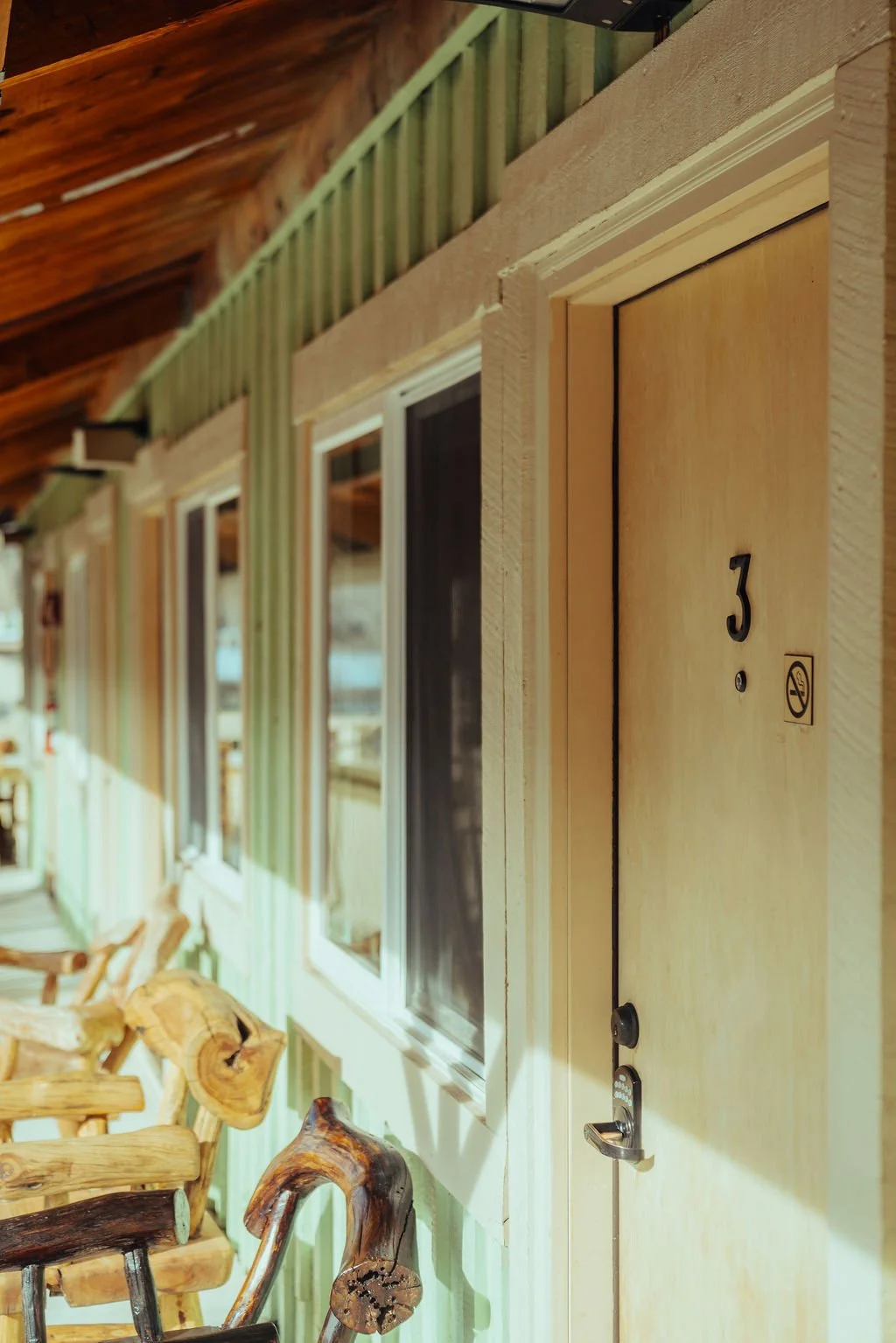 A wooden exterior wall with a door labeled '3' and a no smoking sign. There are multiple windows and wooden chairs along the wall, with a rustic ceiling above.