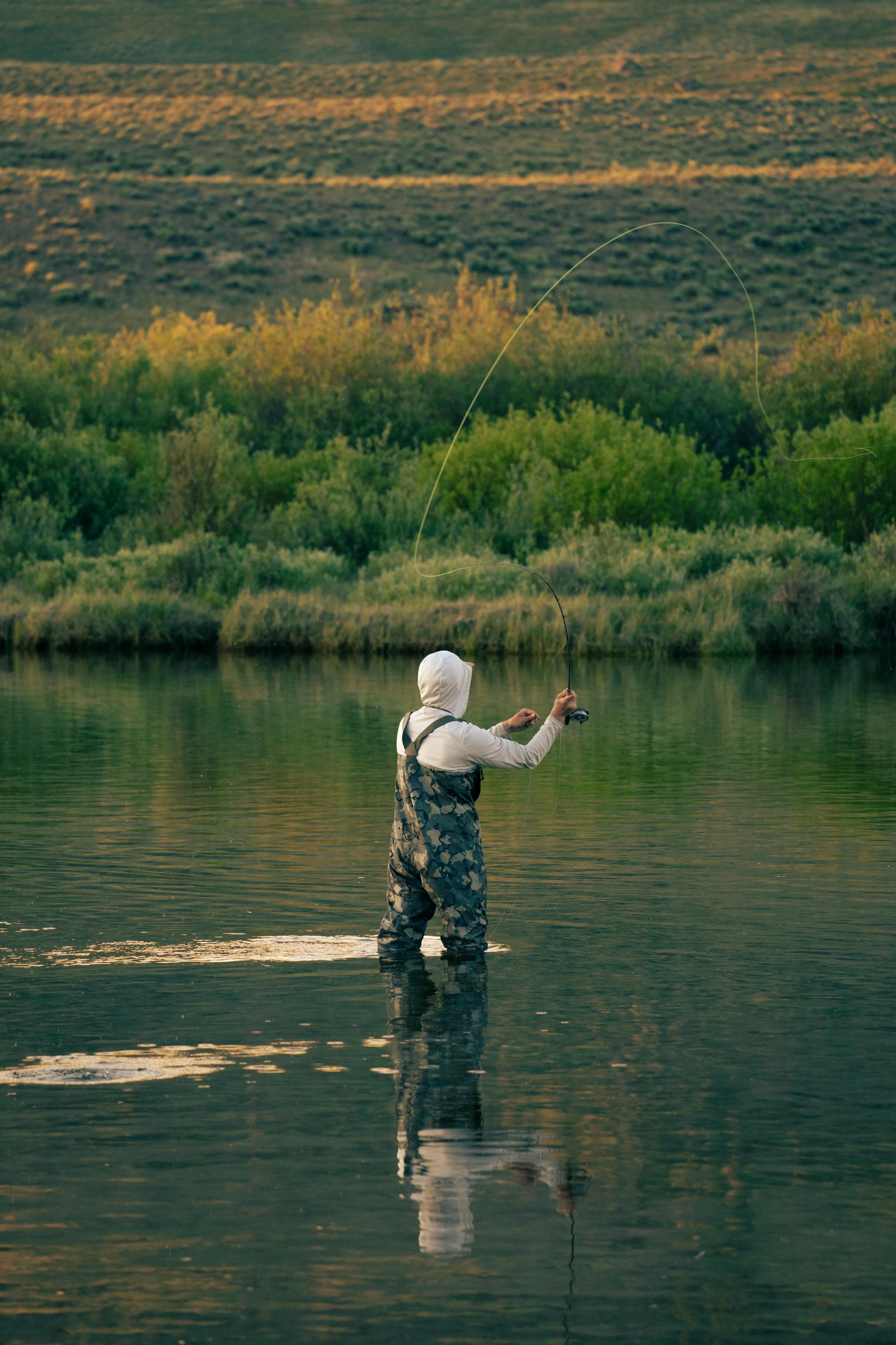 Person fishing in a calm river with lush green trees and open fields in the background during sunset.