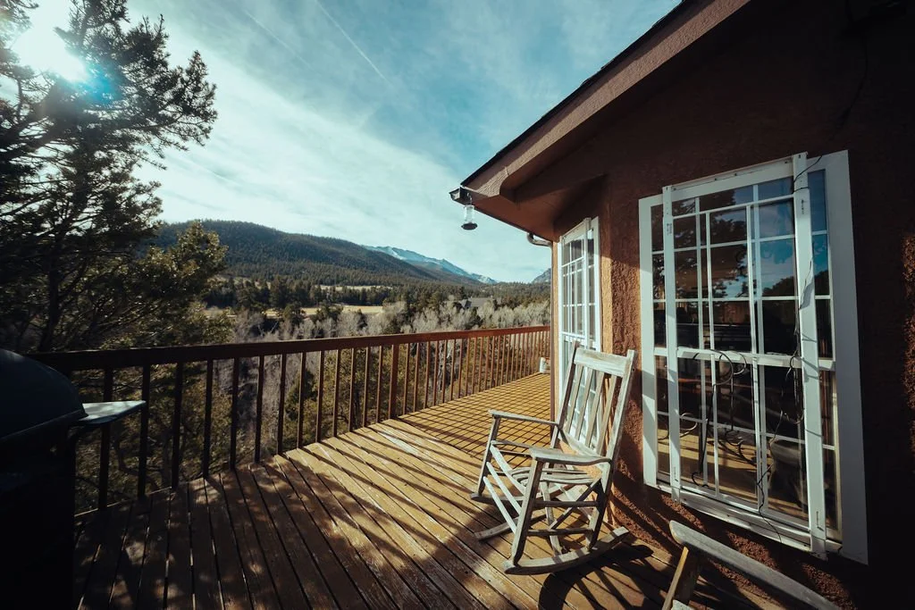 Sunlit wooden balcony with white chairs, overlooking a forested mountain landscape with blue sky and clouds.